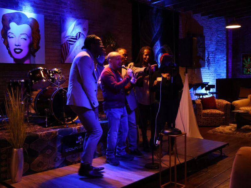 a group of attendees singing together at a microphone on a small stage in a lounge venue with blue and purple stage lighting, drum kit and pop art prints visible in the background
