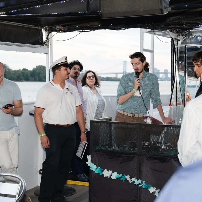 Chris Gittings speaking into a microphone to guests on the deck of the Ben Franklin Yacht, Ben Franklin Bridge in the distance