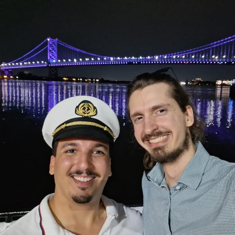 Chris Gittings and a guest in a captain's hat taking a selfie at night with the lit-up Ben Franklin Bridge behind them
