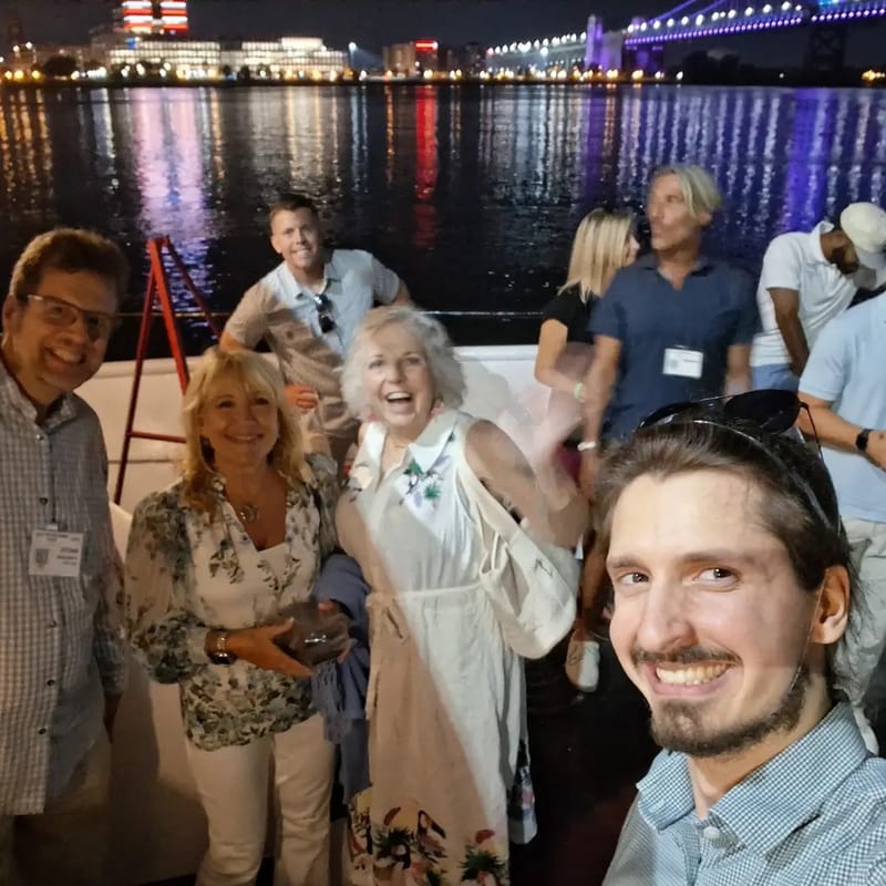 Chris Gittings taking a selfie with smiling guests on the deck at night, city lights on the river behind them
