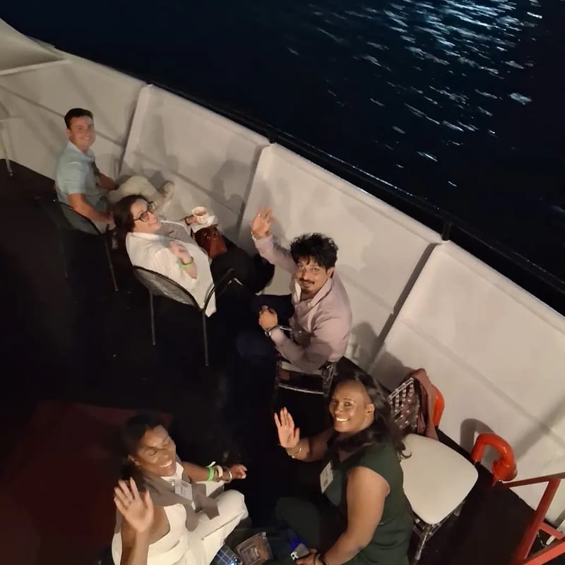 Guests waving up at the camera from a seating area along the side of the yacht at night